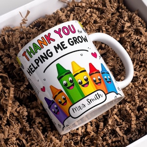 Puede incluir: Taza de cerámica blanca con asa, con el texto "THANK YOU for HELPING ME GROW" en letras coloridas. La taza tiene ilustraciones de crayones sonrientes y el nombre "Mrs Smith". La taza está dentro de una caja blanca con relleno de papel marrón.