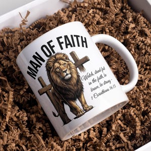 May include: White ceramic mug with the text "MAN OF FAITH" and a detailed illustration of a lion with a cross. The mug also features a Bible verse: "Watch, stand fast in the faith, be brave, be strong - 1 Corinthians 16:13."