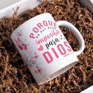 Op de afbeelding: Witte keramische mok met een handvat, met roze tekst en vlindergraphics. De mok heeft de tekst "Porque no hay nada imposible para DIOS" en het bijbelvers "Lucas 1:37". De mok is omgeven door bruin verpakkingsmateriaal.