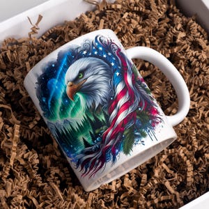 May include: White ceramic mug featuring a detailed illustration of an American bald eagle with a patriotic design. The artwork includes the American flag, a forest, and the Northern Lights. The mug has a white handle and is presented in a gift box with brown packing material.