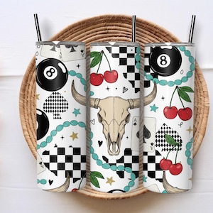 May include: Three white stainless steel tumblers with black straws. The tumblers feature a repeating pattern of an eight ball, cherries, playing cards, a bull skull, and a turquoise beaded chain. The background is a woven basket.