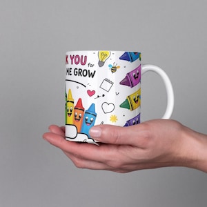 May include: White ceramic mug featuring colorful cartoon crayons with smiling faces. The mug has the text "Thank You for Helping Me Grow" and includes a lightbulb, bee, and other small illustrations. The mug is held in a person's hand.