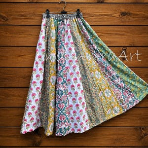 May include: A long, flared skirt featuring a patchwork design of various floral patterns in shades of pink, yellow, green, and white. The waistband is striped in black and white, and the skirt hangs from a metal hanger against a wooden background.