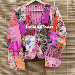 Peut inclure: Veste patchwork avec col en V et fermeture boutonnée. La veste présente une variété de motifs floraux et géométriques dans les tons de rose, d'orange et de vert. Le texte "Indie Aura Art" est visible sur la veste.