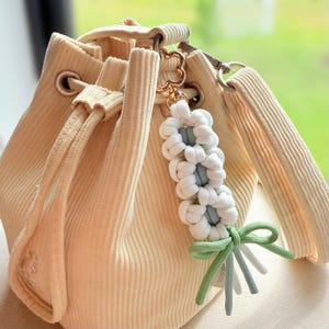 Könnte beinhalten: Cremefarbene Cord-Bucket-Bag mit Kordelzugverschluss. Ein weiß-blaues Makramee-Schlüsselanhänger mit grüner Schleife ist an der Tasche befestigt. Die Tasche hat einen goldfarbenen herzförmigen Verschluss.