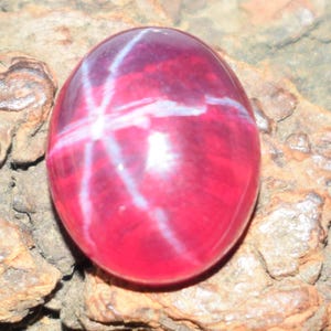 May include: A polished, oval ruby gemstone displaying a six-rayed star effect. The gem is a deep red colour with white star-shaped inclusions. The ruby is set against a rough, textured, brown surface.