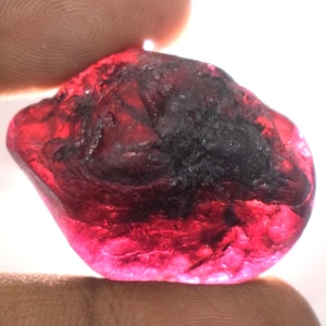 May include: A raw, uncut gemstone, predominantly deep red with dark, almost black inclusions. The stone has a natural, irregular shape and a slightly translucent appearance, with a polished surface.