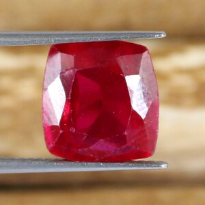 May include: A faceted, cushion-cut ruby gemstone held by tweezers. The ruby is a deep, saturated red color with multiple facets that reflect light. The gemstone is held against a blurred, light brown background.