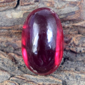 Könnte beinhalten: Ein polierter, oval geformter, tiefroter Edelstein. Der Edelstein hat eine glatte, reflektierende Oberfläche mit Glanzlichtern und Schatten, die Tiefe suggerieren. Der Stein ist vor einem strukturierten, braunen Hintergrund platziert, was seine satte Farbe und seinen Glanz verstärkt. Dieser Edelstein ist für Schmuck geeignet.
