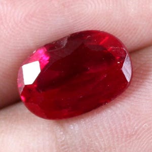 May include: A faceted, oval-shaped ruby gemstone. The gem is a deep, saturated red colour with multiple facets that reflect light. The ruby is set against a neutral background.