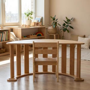 May include: A light-colored wooden children's table and chair set. The table has a curved base and vertical supports. The chair has a ladder back design. The set is in a bright, naturally lit room with other wooden furniture.