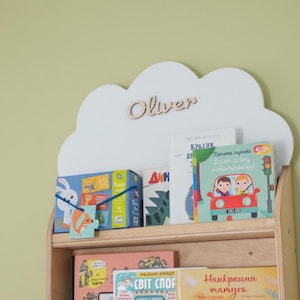 Pode incluir: Uma estante de madeira com um topo em forma de nuvem, com o nome "Oliver" em letras de madeira. A estante contém vários livros infantis e um quebra-cabeça. O fundo é uma parede verde clara.