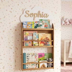 Op de afbeelding: Houten boekenplank met drie planken, gevuld met kleurrijke kinderboeken. De boekenplank is aan een muur met een bloemenpatroon bevestigd. Een wolk-vormig bord met de naam "Sophia" staat boven de planken. Een knuffelolifant staat op de onderste plank.