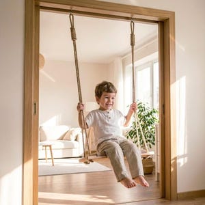 May include: A child is seated on a wooden swing with rope supports, suspended from a doorway. The swing is made of light-colored wood and natural rope. The child is wearing a white shirt and tan pants, smiling. The background includes a living room with a couch and a plant.