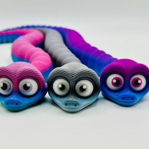 May include: Three toy snakes in shades of blue, pink, purple, and grey, each with large, cartoonish eyes. The heads have textured scales and expressive eyes. The snakes are designed to be playful and colourful.