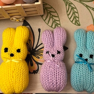May include: Five knitted bunny plushes in different colours: teal, yellow, purple, light blue, and pink. Each bunny has a small bow tied around its neck.