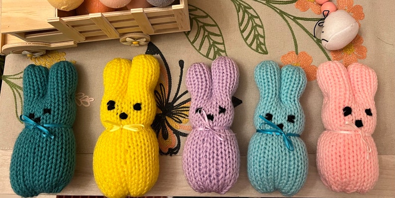 May include: Five knitted bunny plushes in different colours: teal, yellow, purple, light blue, and pink. Each bunny has a small bow tied around its neck.