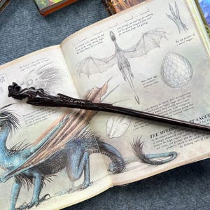 以下が含まれることがあります： 精巧に彫刻された濃い茶色の杖が、ドラゴンなどの生き物のイラストが描かれた開いた本の上に置かれています。本には、これらの生き物に関する詳細な図とテキストが掲載されています。
