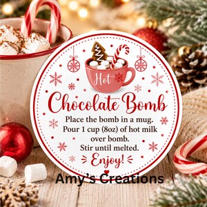 May include: A round white sticker with a red border featuring a red mug illustration with marshmallows, a candy cane, and a gingerbread tree. Text reads: Hot Chocolate Bomb, Place the bomb in a mug. Pour 1 cup (8oz) of hot milk over bomb. Stir until melted. Enjoy! Amy's Creations.