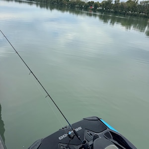 Puede incluir: Una ca&ntilde;a de pescar est&aacute; montada en una moto acu&aacute;tica negra y azul, con el agua al fondo. El agua est&aacute; tranquila y refleja el cielo nublado y los &aacute;rboles de la orilla lejana.
