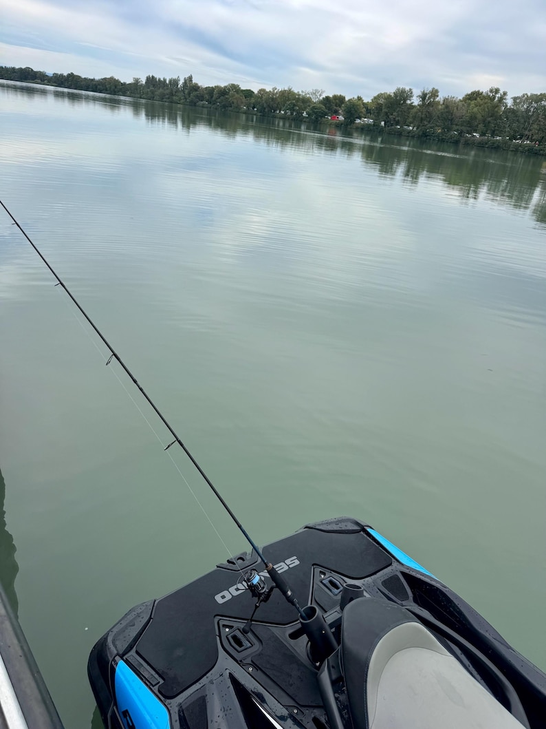Puede incluir: Una ca&ntilde;a de pescar est&aacute; montada en una moto acu&aacute;tica negra y azul, con el agua al fondo. El agua est&aacute; tranquila y refleja el cielo nublado y los &aacute;rboles de la orilla lejana.