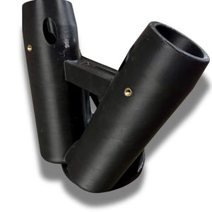 Puede incluir: Un objeto negro impreso en 3D con dos aberturas cil&iacute;ndricas conectadas por un puente rectangular. Cada cilindro tiene un peque&ntilde;o agujero circular y un remache color lat&oacute;n. El objeto tiene una forma curva y est&aacute; sobre un fondo blanco.