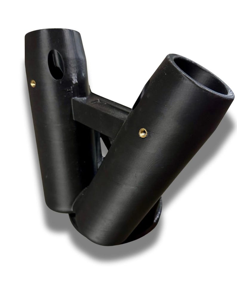 Puede incluir: Un objeto negro impreso en 3D con dos aberturas cil&iacute;ndricas conectadas por un puente rectangular. Cada cilindro tiene un peque&ntilde;o agujero circular y un remache color lat&oacute;n. El objeto tiene una forma curva y est&aacute; sobre un fondo blanco.