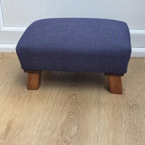 May include: A rectangular footstool featuring a textured, dark blue fabric upholstery with a subtle woven pattern. The seat is supported by four short, light brown wooden legs with a visible grain, providing a stable base for the cushioned top.
