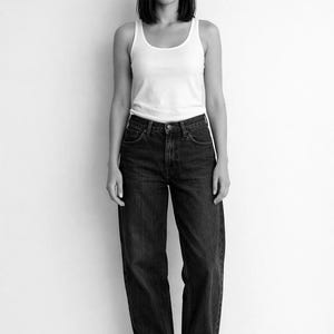 Könnte beinhalten: Schwarz-Weiß-Foto einer Person in einem weißen Tanktop und einer dunkelblauen Jeans. Die Jeans hat eine lockere Passform. Die Person steht vor einer weißen Wand.