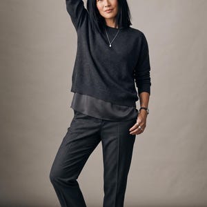 May include: A dark charcoal gray long-sleeved sweater with a round neckline layered over a dark gray satin-like camisole, paired with matching dark charcoal gray tailored trousers. The outfit includes a silver necklace with a small pendant.