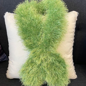May include: A green fluffy scarf made of yarn, displayed on a white pillow on a dark gray couch.