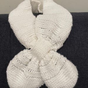 May include: A white knitted scarf with a knotted design. The scarf is made of a soft, textured yarn and has a loose, flowing shape.