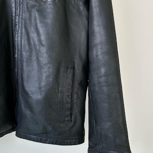 May include: A black leather jacket featuring a full-length front zipper, a high collar, and two vertical slit pockets on the lower front. The jacket has long sleeves and a smooth, textured finish across the entire surface of the garment.