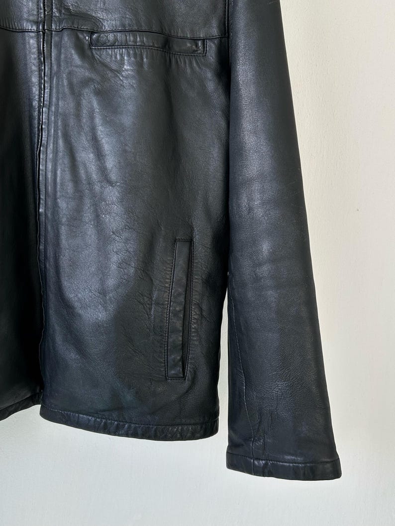 May include: A black leather jacket featuring a full-length front zipper, a high collar, and two vertical slit pockets on the lower front. The jacket has long sleeves and a smooth, textured finish across the entire surface of the garment.