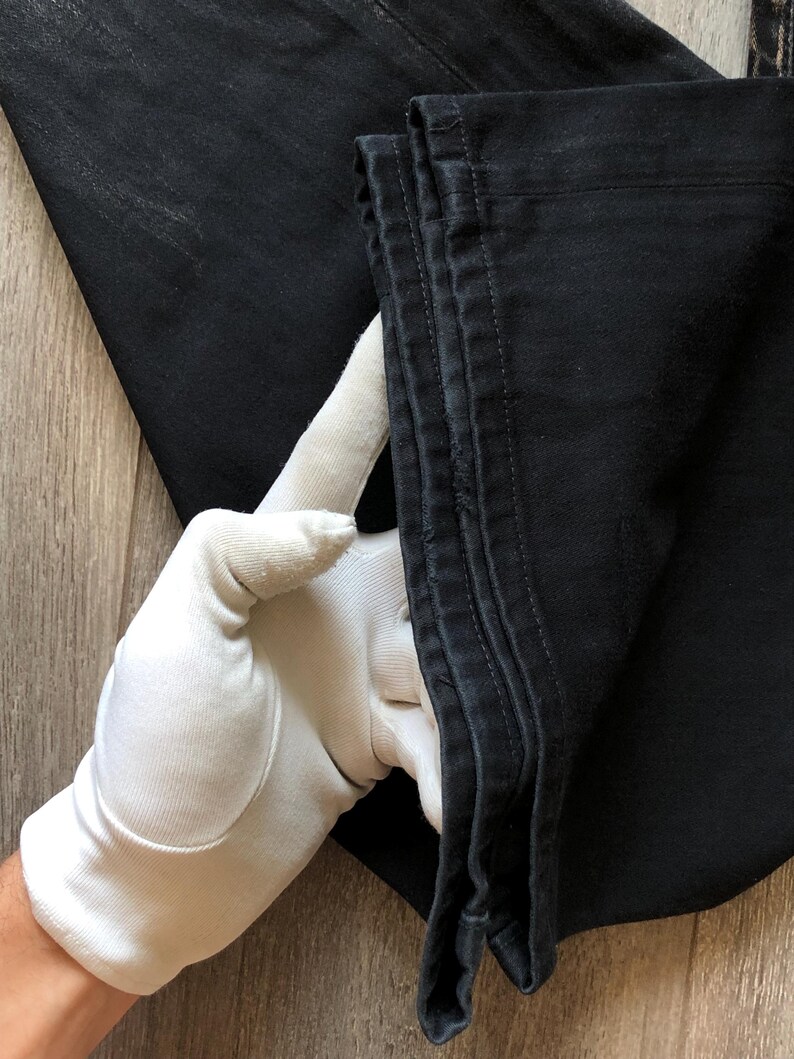 Puede incluir: Primer plano de unos vaqueros de mezclilla negros. Los vaqueros est&aacute;n doblados, mostrando las costuras y la textura de la tela. Un guante blanco sostiene el borde doblado, resaltando el color oscuro y los detalles de las costuras.