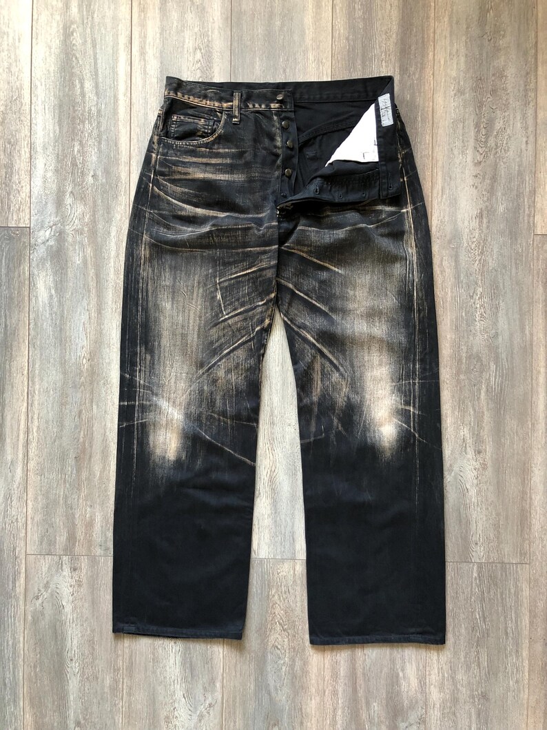 Puede incluir: Vaqueros de mezclilla negros con un aspecto descolorido y desgastado. Los vaqueros tienen una bragueta con botones y un dise&ntilde;o cl&aacute;sico de cinco bolsillos. La tela muestra patrones de desgaste marr&oacute;n claro y blanco, creando un aspecto vintage. Los vaqueros est&aacute;n planos sobre una superficie de madera.
