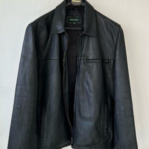 May include: A black leather jacket with a classic collar, a full-length front zipper, and two side pockets. The interior label displays the brand name Springfield. The jacket is hung on a black hanger against a plain, light-colored wall background.