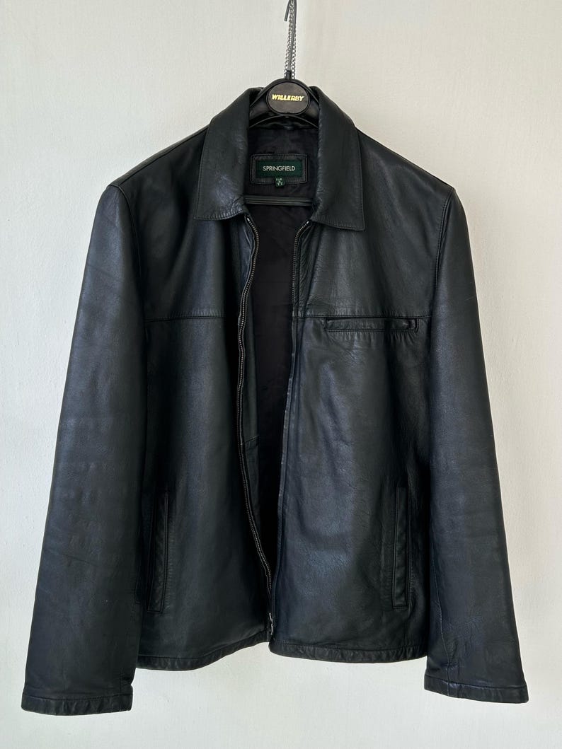 May include: A black leather jacket with a classic collar, a full-length front zipper, and two side pockets. The interior label displays the brand name Springfield. The jacket is hung on a black hanger against a plain, light-colored wall background.