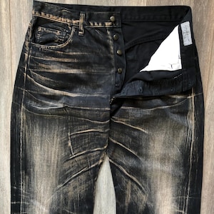 Puede incluir: Vaqueros de mezclilla negros con un dise&ntilde;o desgastado y descolorido. Los vaqueros tienen bragueta con botones y un dise&ntilde;o cl&aacute;sico de cinco bolsillos. La tela tiene un aspecto desgastado y texturizado con acentos marr&oacute;n claro.