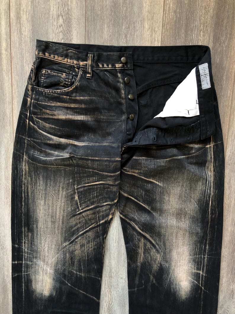 Puede incluir: Vaqueros de mezclilla negros con un dise&ntilde;o desgastado y descolorido. Los vaqueros tienen bragueta con botones y un dise&ntilde;o cl&aacute;sico de cinco bolsillos. La tela tiene un aspecto desgastado y texturizado con acentos marr&oacute;n claro.