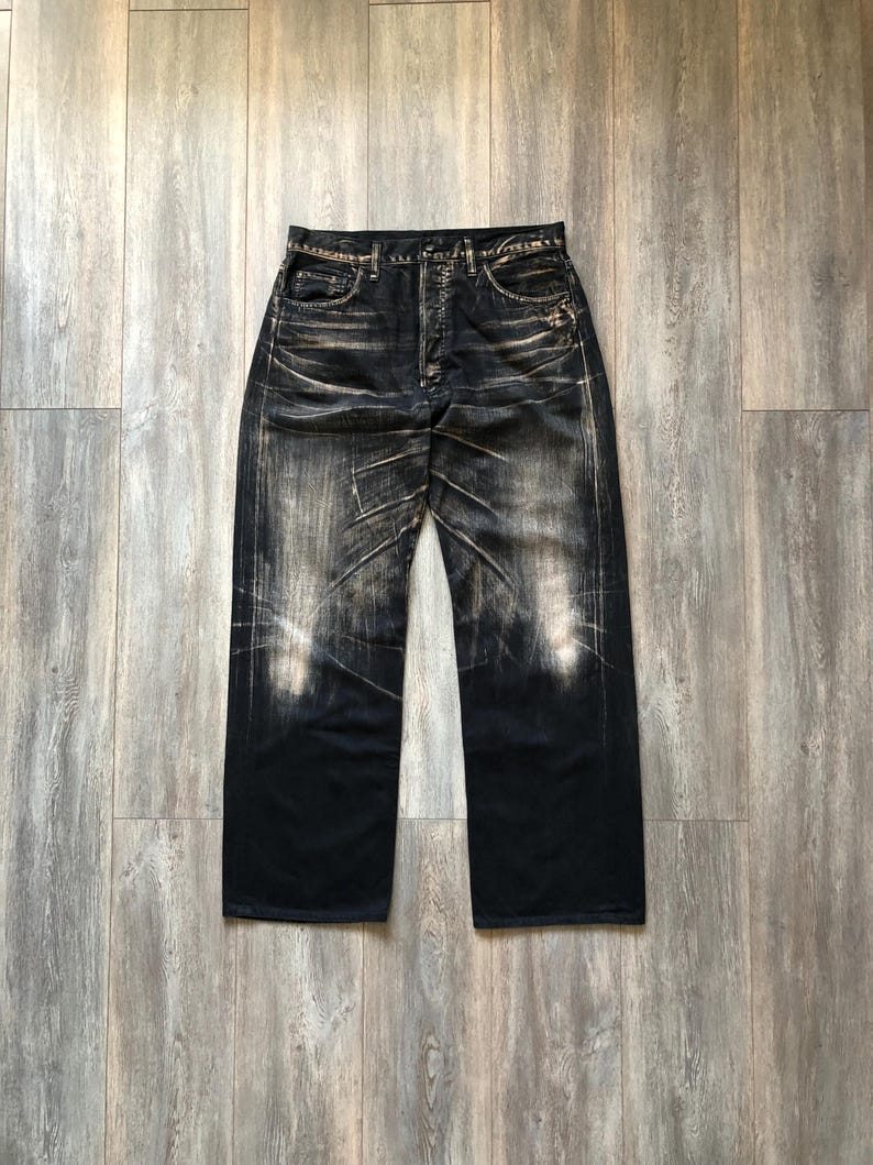 Puede incluir: Vaqueros de mezclilla de lavado oscuro con un dise&ntilde;o desgastado y descolorido. Los vaqueros tienen un dise&ntilde;o cl&aacute;sico de cinco bolsillos y un corte de pierna recta. La mezclilla tiene una apariencia desgastada con &aacute;reas m&aacute;s claras y marcas de desgaste visibles.