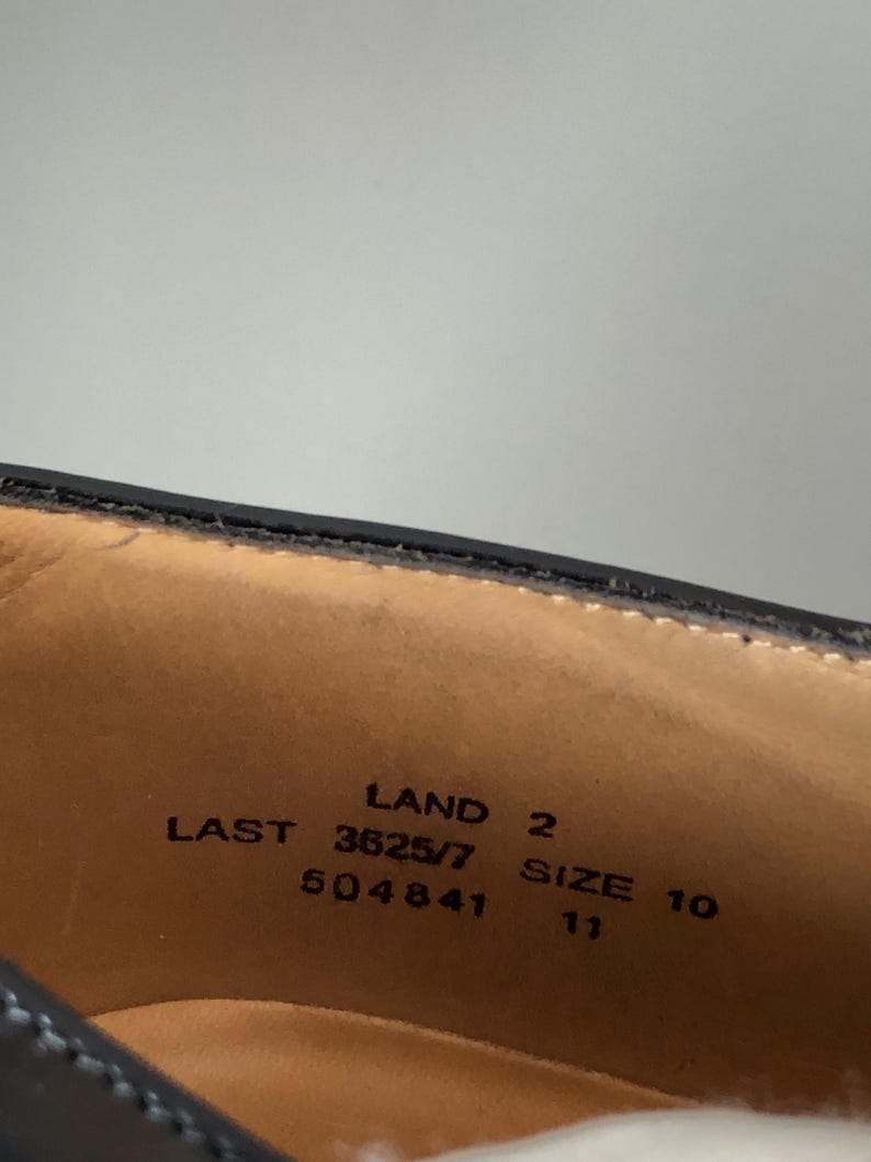 May include: Close-up of the inside of a black leather shoe. The shoe's interior is tan with black text that reads "LAND 2", "LAST 3625/7", "SIZE 10", and "504841". The size is also listed as 11.