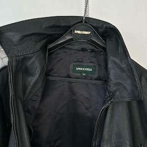 May include: A black leather jacket with a pointed collar, a front zipper closure, and a zippered pocket on the lower right side. The interior features a black lining and a rectangular label with the brand name SPRINGFIELD in white text.
