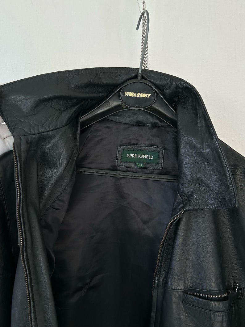 May include: A black leather jacket with a pointed collar, a front zipper closure, and a zippered pocket on the lower right side. The interior features a black lining and a rectangular label with the brand name SPRINGFIELD in white text.