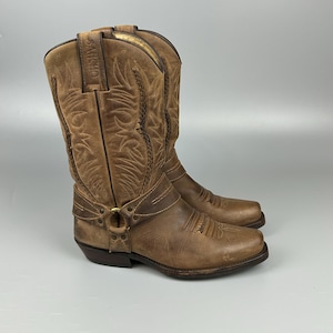 May include: A pair of brown leather cowboy boots with intricate stitching and a harness detail. The boots have a worn, distressed look, with the word "SANCHO" embossed on the top. The boots are approximately 12 inches tall.