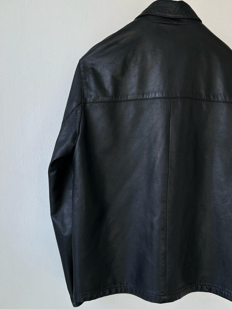 May include: A black leather jacket featuring a classic collar, long sleeves, and a horizontal back yoke seam. The garment displays a smooth, matte texture with visible stitching along the seams and a straight hemline, presented from a rear view.