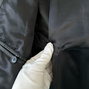 May include: A black jacket interior featuring a smooth satin lining, a single black button attached to the leather trim, and a small area of visible stitching or repair on the fabric panel near the center seam.
