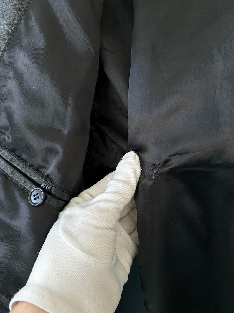May include: A black jacket interior featuring a smooth satin lining, a single black button attached to the leather trim, and a small area of visible stitching or repair on the fabric panel near the center seam.