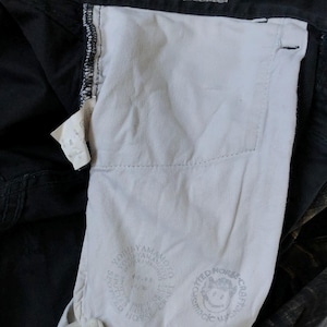 Puede incluir: Primer plano de pantalones negros con forro de bolsillo blanco. El forro presenta un logotipo circular con texto y un gr&aacute;fico de caballo sonriente. Una peque&ntilde;a etiqueta con texto es visible cerca del bolsillo. Los pantalones est&aacute;n doblados, mostrando el interior.