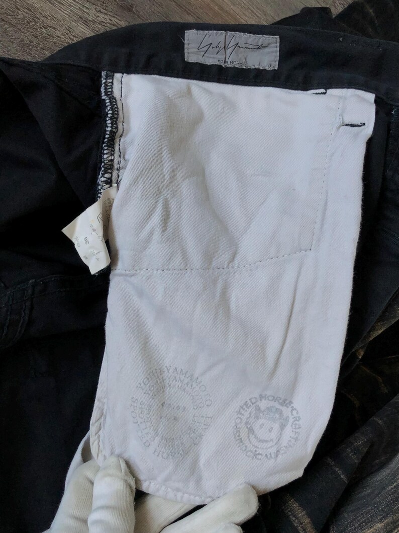 Puede incluir: Primer plano de pantalones negros con forro de bolsillo blanco. El forro presenta un logotipo circular con texto y un gr&aacute;fico de caballo sonriente. Una peque&ntilde;a etiqueta con texto es visible cerca del bolsillo. Los pantalones est&aacute;n doblados, mostrando el interior.
